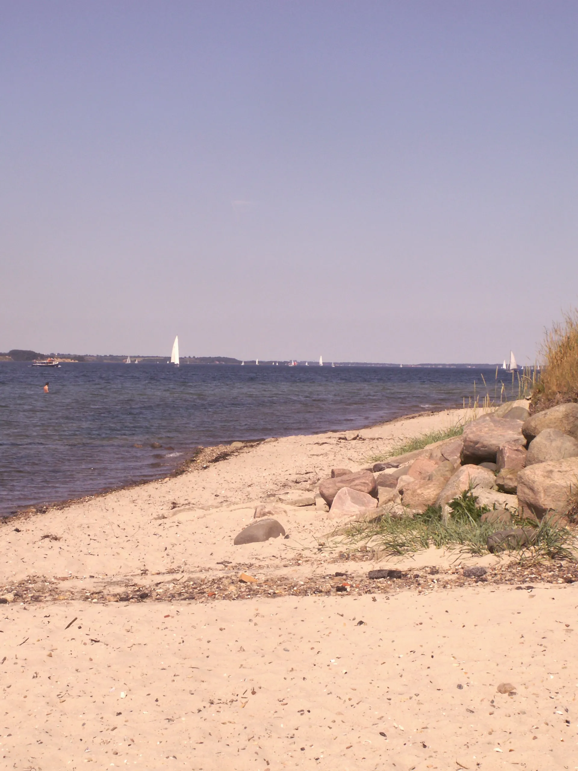 Sandstrand mit Findlingen und Strandgras, im Hintergrund Segelboote